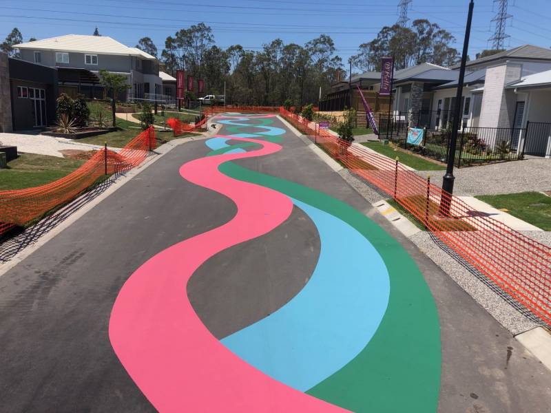 A suburban road transformed with decorative asphalt sealant in pink, green, and blue waves as part of a streetscaping project.