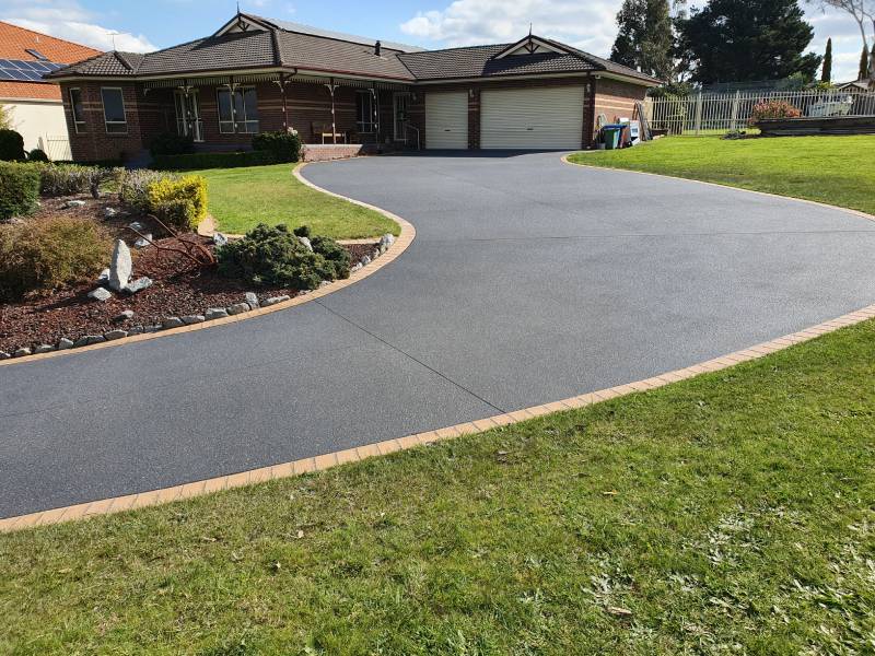 A freshly resurfaced, curved concrete driveway with a textured dark finish and contrasting brick edging in front of a suburban home.