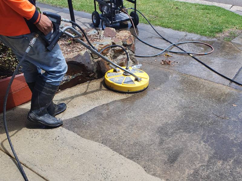 A technician uses a surface cleaner attachment on a pressure washer to grind and clean a concrete driveway before sealing.