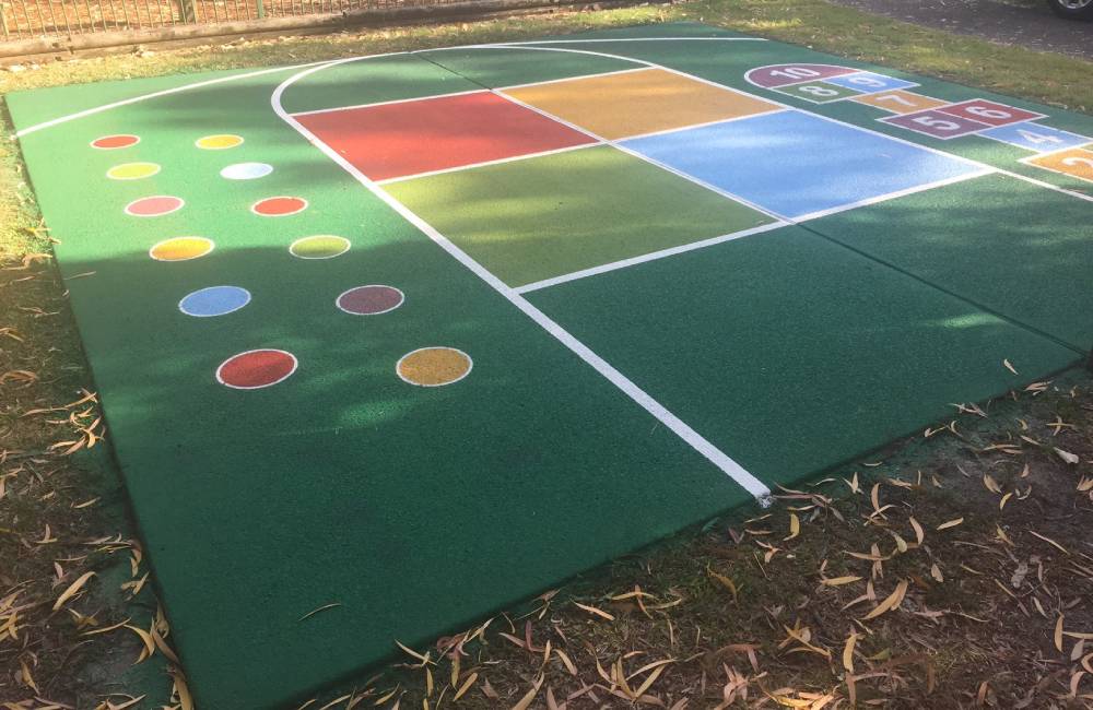 A brightly coloured outdoor play area with sport flooring featuring hopscotch, circles, and down-ball squares in primary colours.