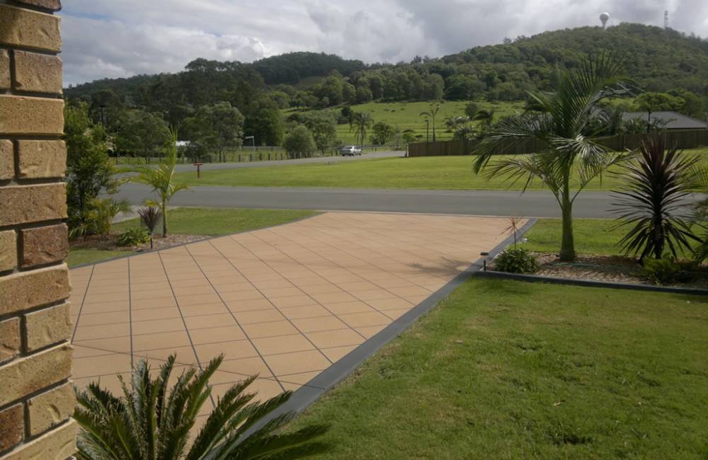 A neat, tan-coloured paved-look driveway leading to a suburban street surrounded by greenery.