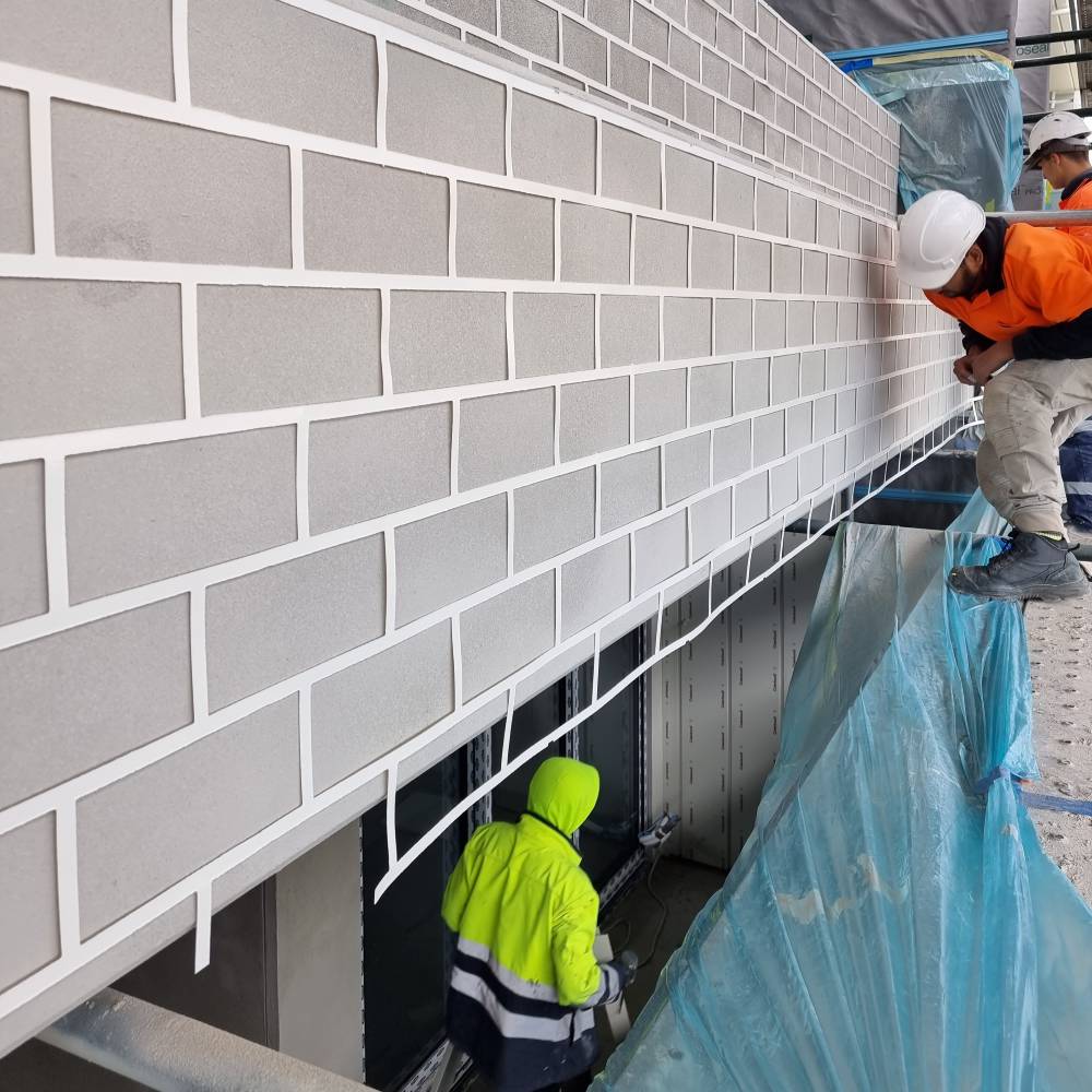 Workers preparing a grey stencil design resembling brickwork on a building facade using tape and masking film.