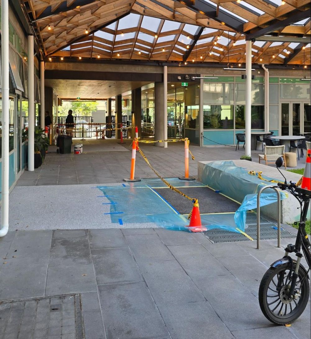 Fresh asphalt resurfacing underway beneath a timber-roofed commercial entryway, with taped-off areas, traffic cones, and construction coverings for safety.