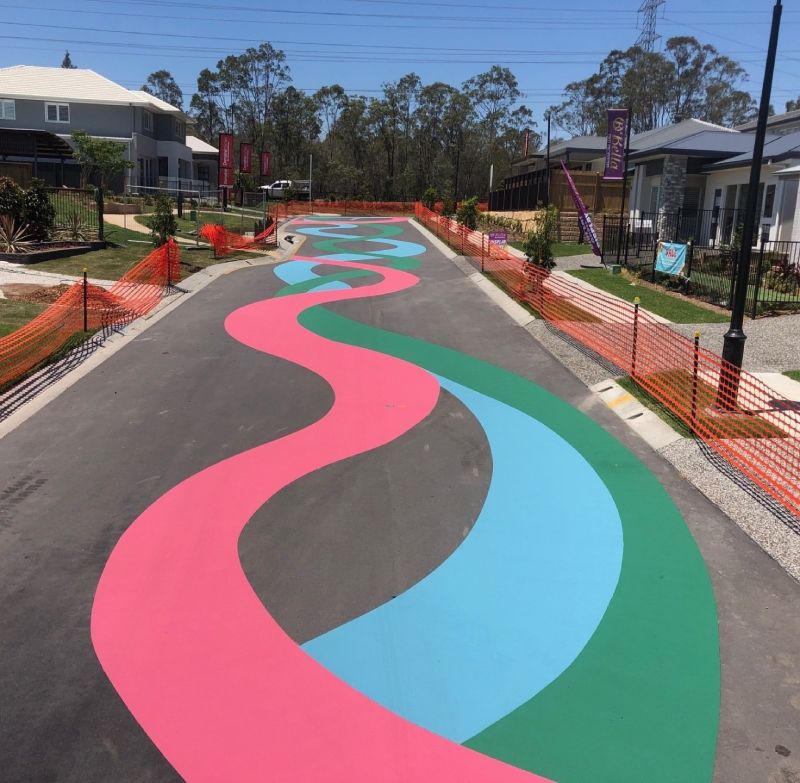 Residential street painted with wide flowing bands of pink, blue, and green asphalt coating, adding playful character to a suburban display village.