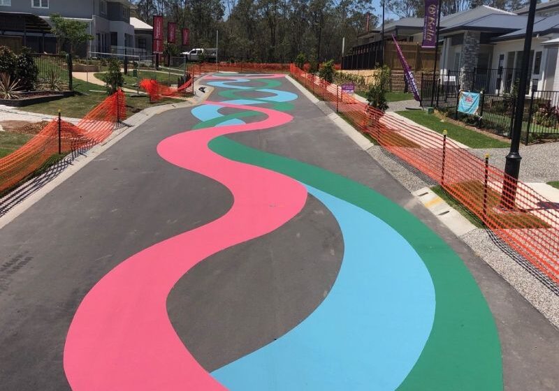 Residential street painted with wide flowing bands of pink, blue, and green asphalt coating, adding playful character to a suburban display village.